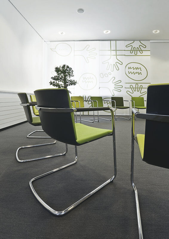 Ergonomische Bürostühle mit grüner Polsterung in einem modern gestalteten Meetingraum mit Wandgrafiken.