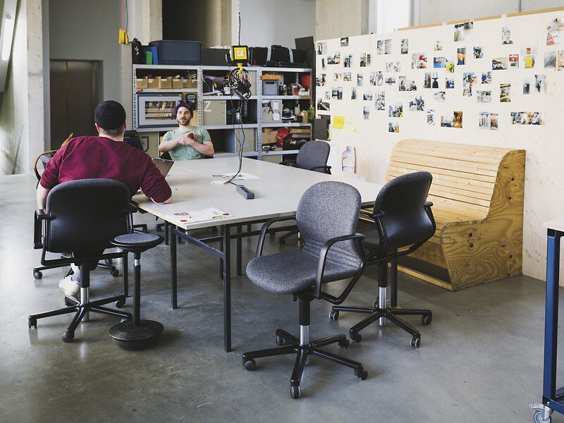 Zwei Männer sitzen in einem modernen Büro mit ergonomischen Stühlen, Tisch und Pinnwand voller Fotos.