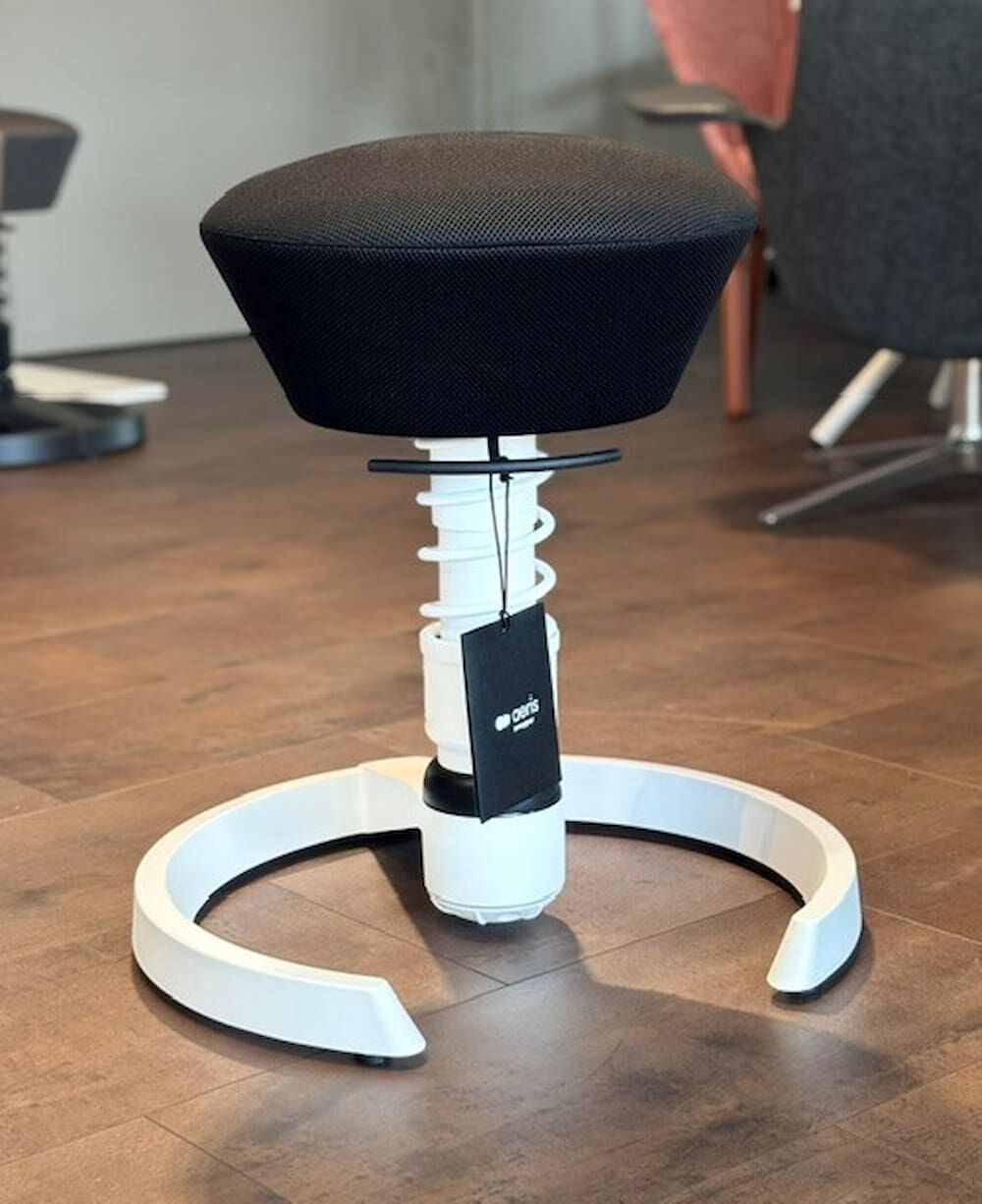 Moderner ergonomischer Hocker mit schwarzem Sitzpolster und weißem Gestell auf Holzboden im Büro.