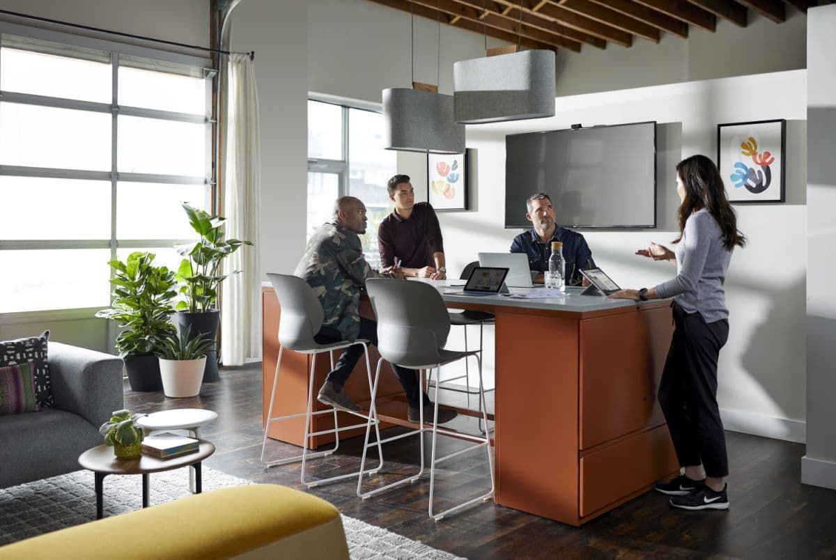 Vier Personen in modernem Büro, sprechen an Stehtisch mit Laptops. Pflanzen und Sofa im Hintergrund.