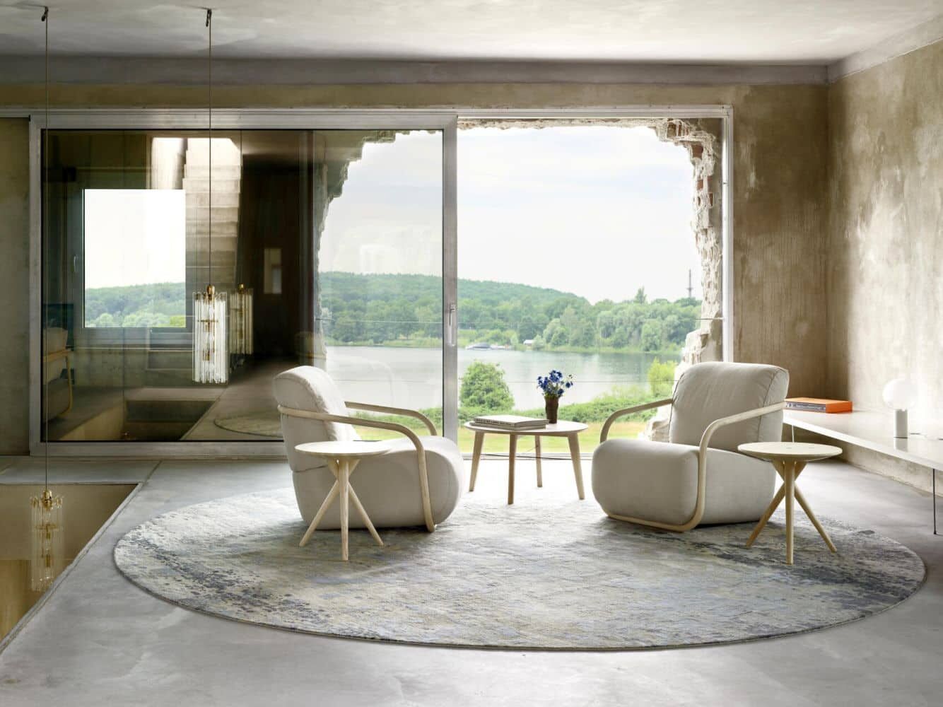 Modern eingerichteter Raum mit zwei Sesseln, Beistelltischen auf rundem Teppich, große Fenster mit Seeblick.