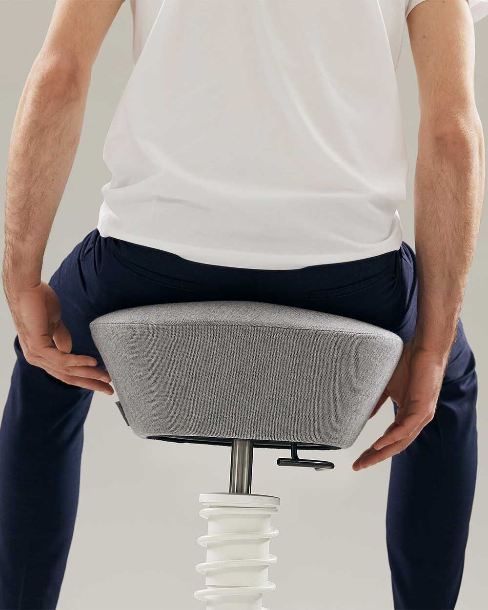 Person sitzt auf einem ergonomischen Hocker mit grauer Polsterung und einem flexiblen, weißen Federfuß.