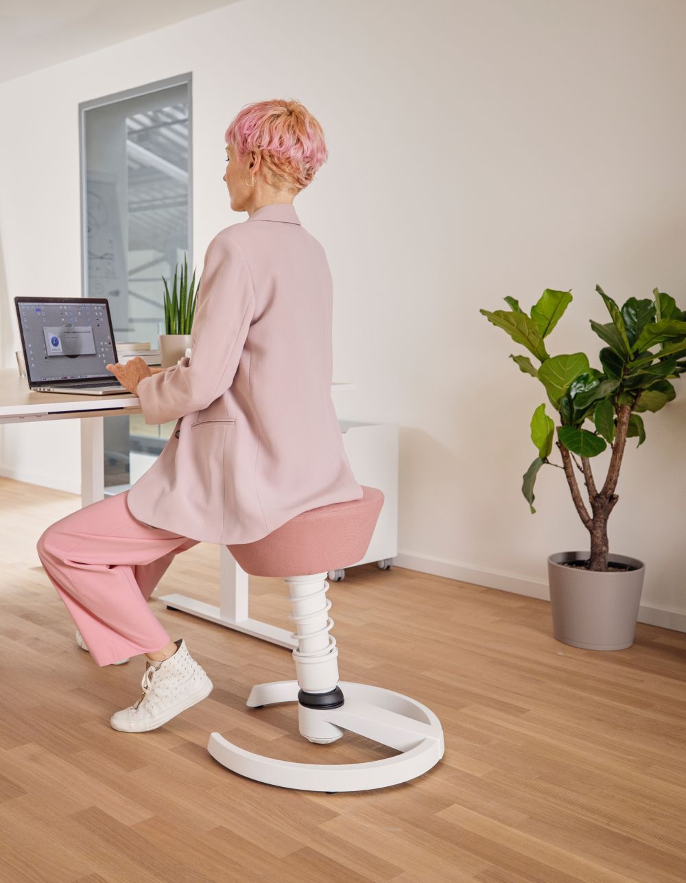Person mit rosa Haaren und Anzug sitzt auf ergonomischem Hocker an einem Schreibtisch mit Laptop.