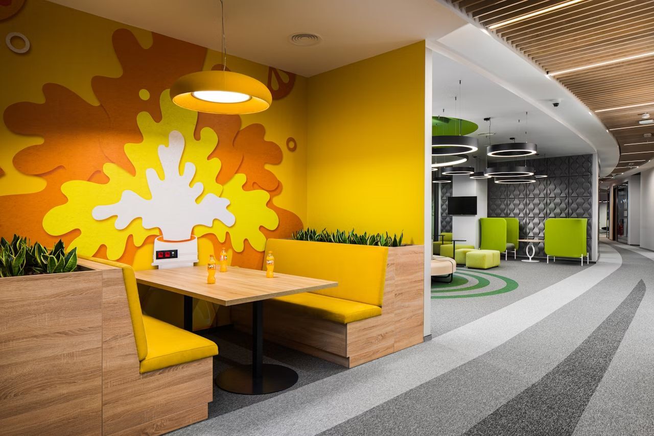Moderne Bürolounge mit gelben Sitzgelegenheiten, leuchtendem Wandbild und grünen Pflanzen für dynamisches Arbeiten.