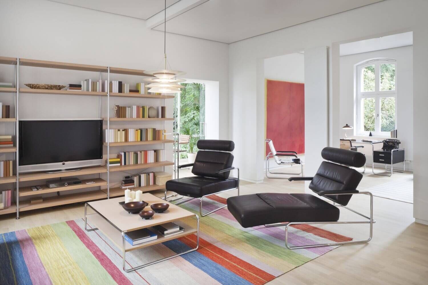 Modernes Wohnzimmer mit ergonomischen Ledersesseln, farbenfrohem Teppich, Bücherregal und Blick ins helle Arbeitszimmer.