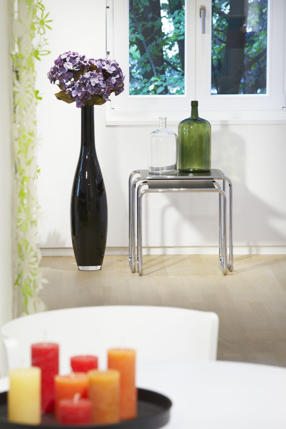 Elegantes Interieur mit schwarzer Vase, lila Blumen, gestapelten Tischen und Vasen vor einem Fenster.