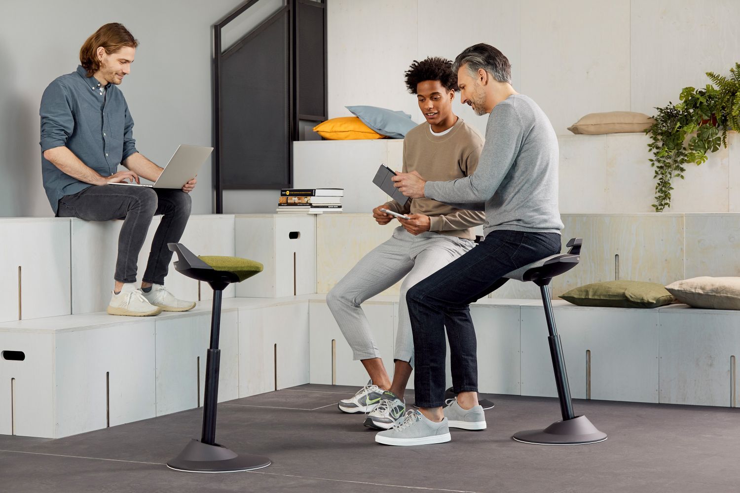 Drei Männer interagieren mit Laptops und Tablets auf ergonomischen Stühlen in einem modernen Bürosetting.