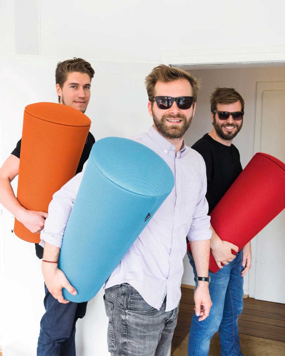 Drei Männer mit Sonnenbrillen halten bunte ergonomische Rückenstützen in blau, rot und orange.