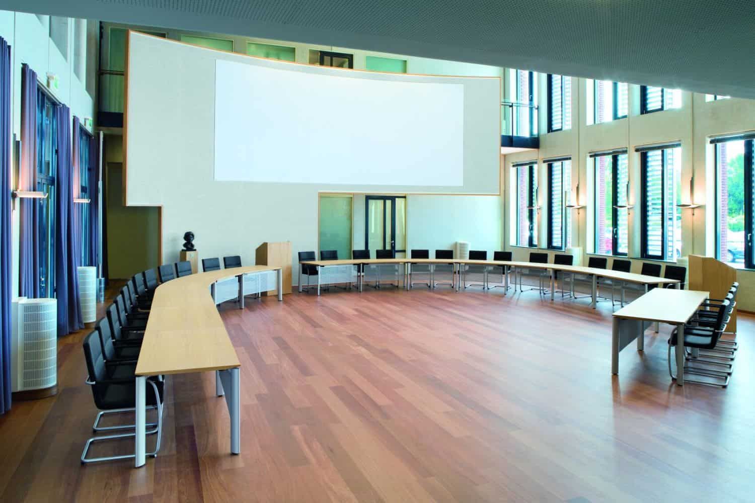 Moderner Konferenzraum mit gebogener Tischanordnung und ergonomischen Stühlen, großer Projektionsfläche und Holzfußboden.