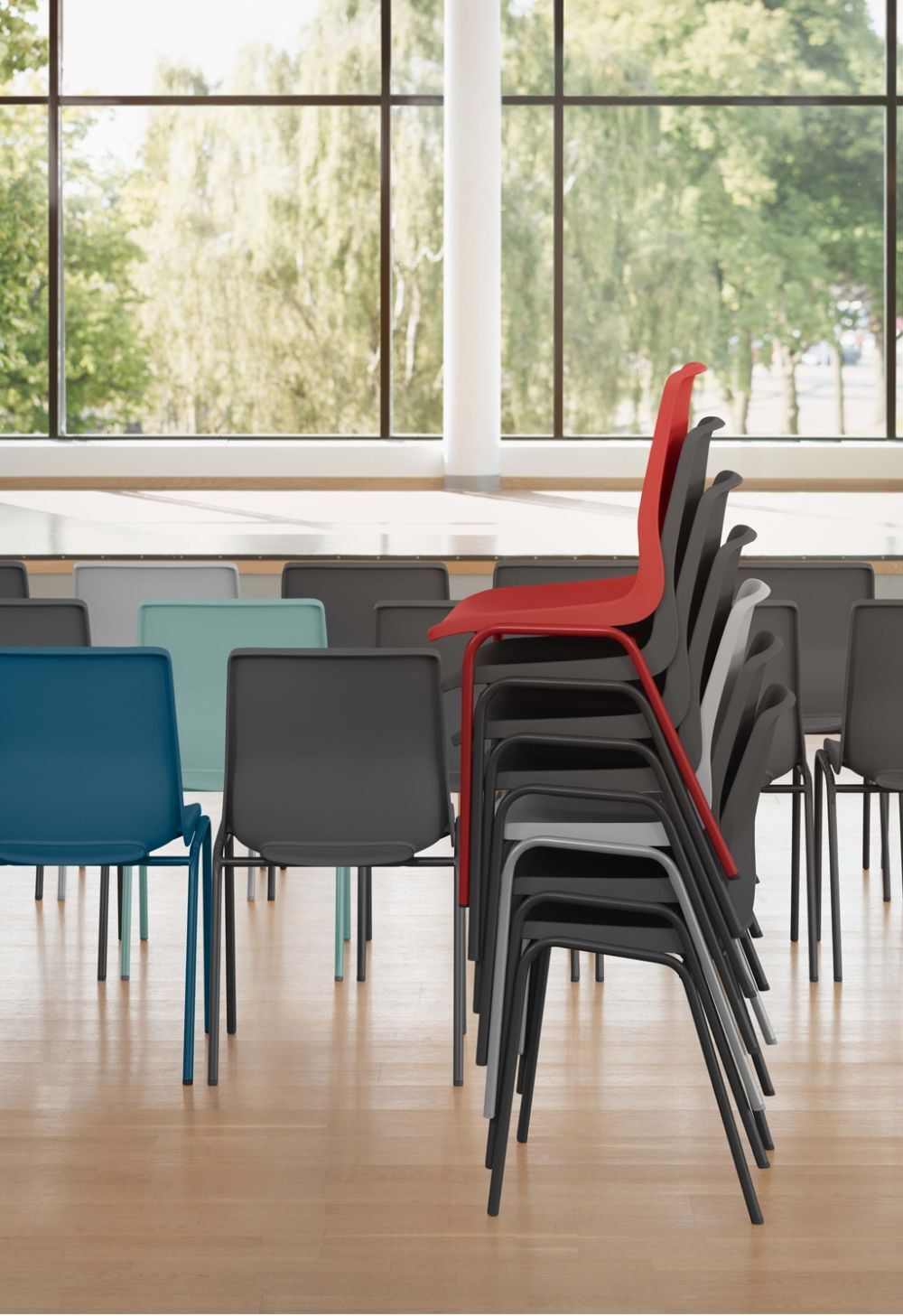 Mehrere gestapelte Stühle in verschiedenen Farben, darunter ein auffälliger roter Stuhl, in einem Raum mit großen Fenstern.