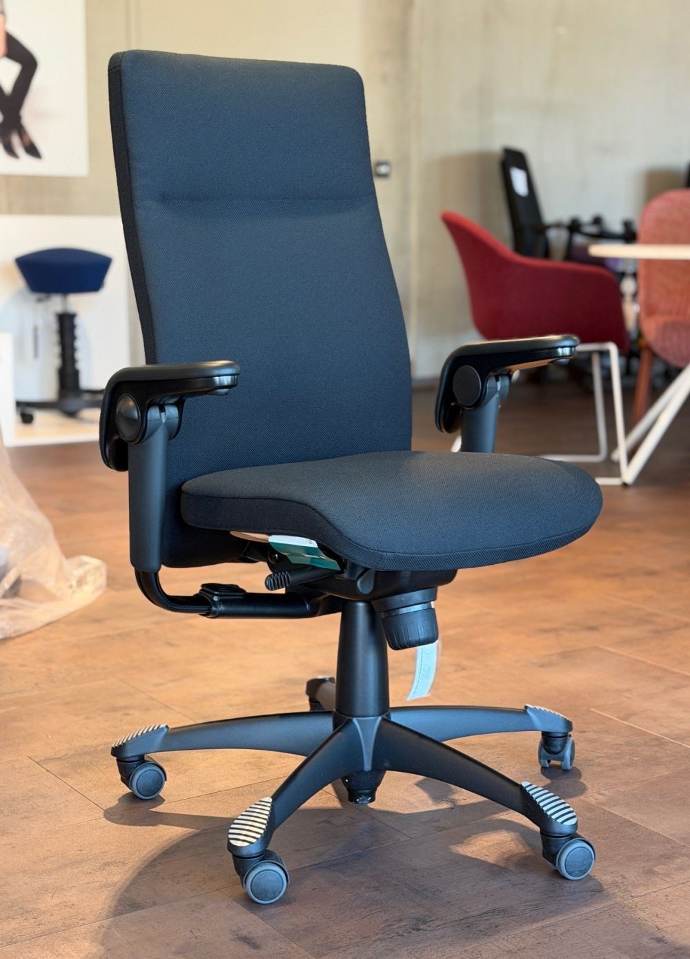 Ergonomischer Bürostuhl mit schwarzem Stoffbezug, verstellbaren Armlehnen und Rollen auf einem Holzfußboden.