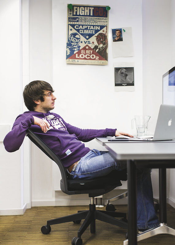 Mann in lila Kapuzenpullover lehnt sich auf ergonomischem Bürostuhl zurück, arbeitet am Laptop, Poster im Hintergrund.