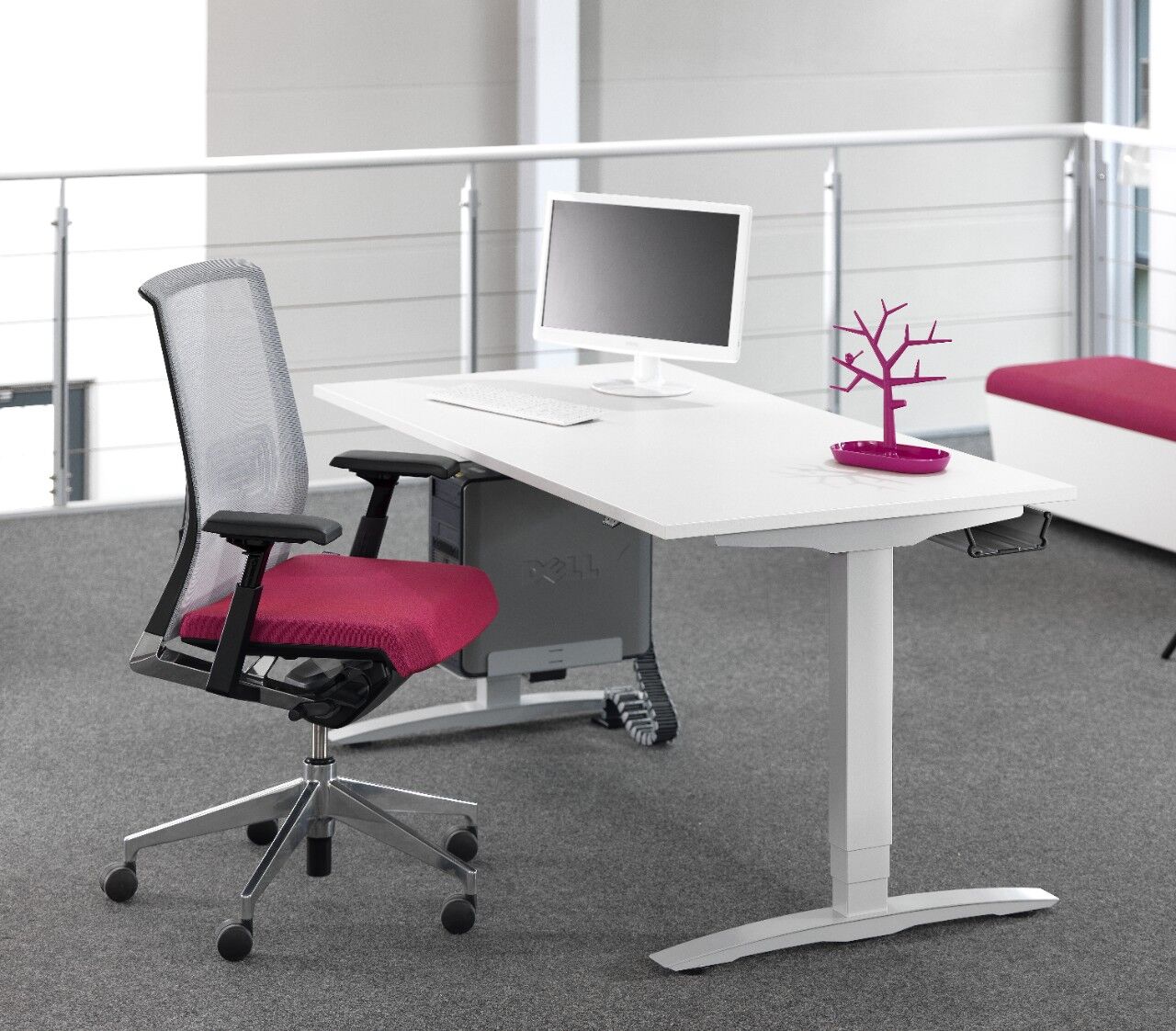 Ergonomischer Schreibtischstuhl mit rotem Sitz an einem weißen Schreibtisch, Monitor und lila Deko auf grauem Teppich.