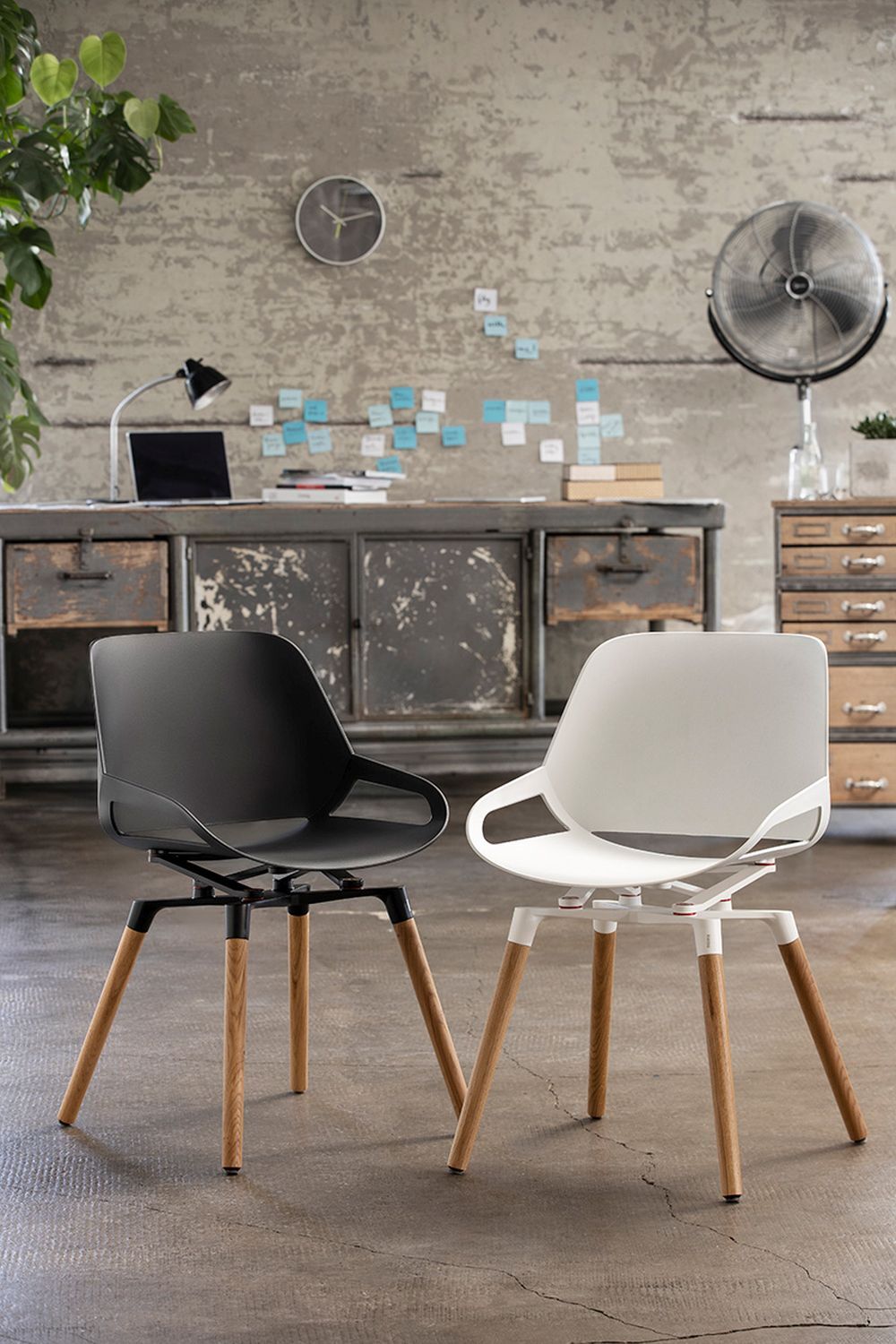 Zwei ergonomische Designerstühle, schwarz und weiß, vor einem industriellen Büroumfeld mit Holztisch und Pflanzen.