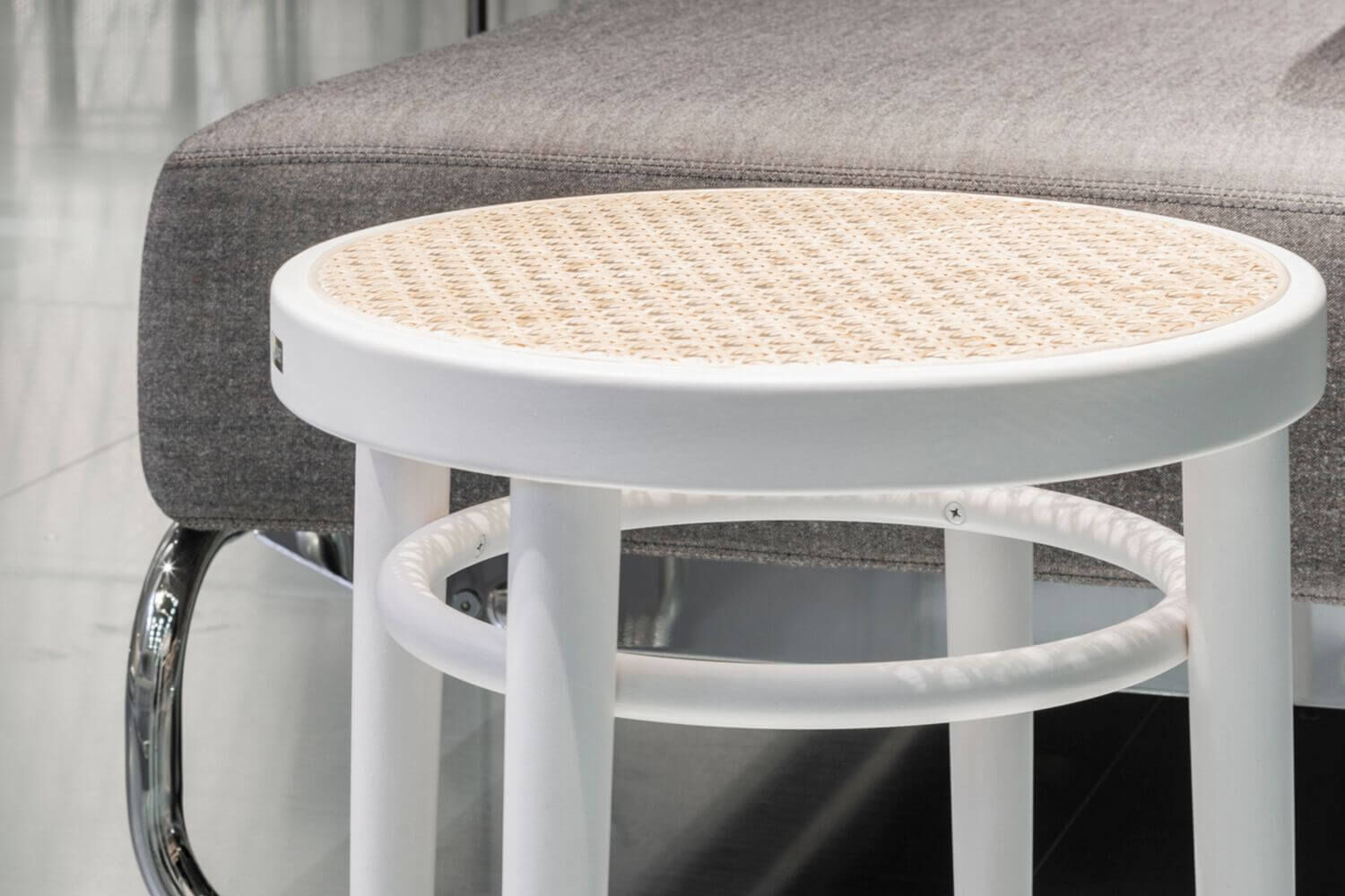 Weißer Hocker mit geflochtener Sitzfläche vor einem grauen Sofateil. Modernes und schlichtes Design.