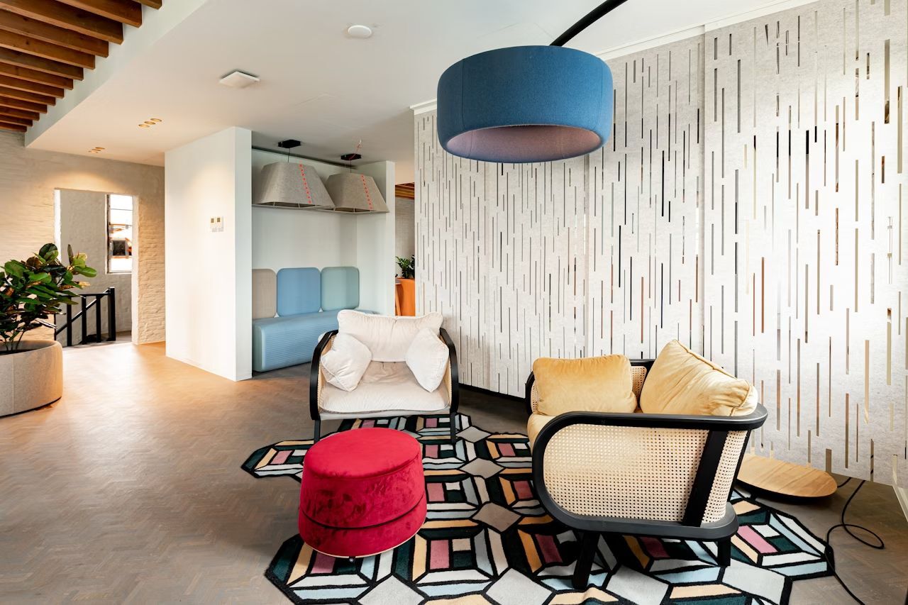 Modernes Wohnzimmer mit stilvollen Sesseln, einem roten Hocker und geometrischem Teppich vor einer strukturierten Wand.