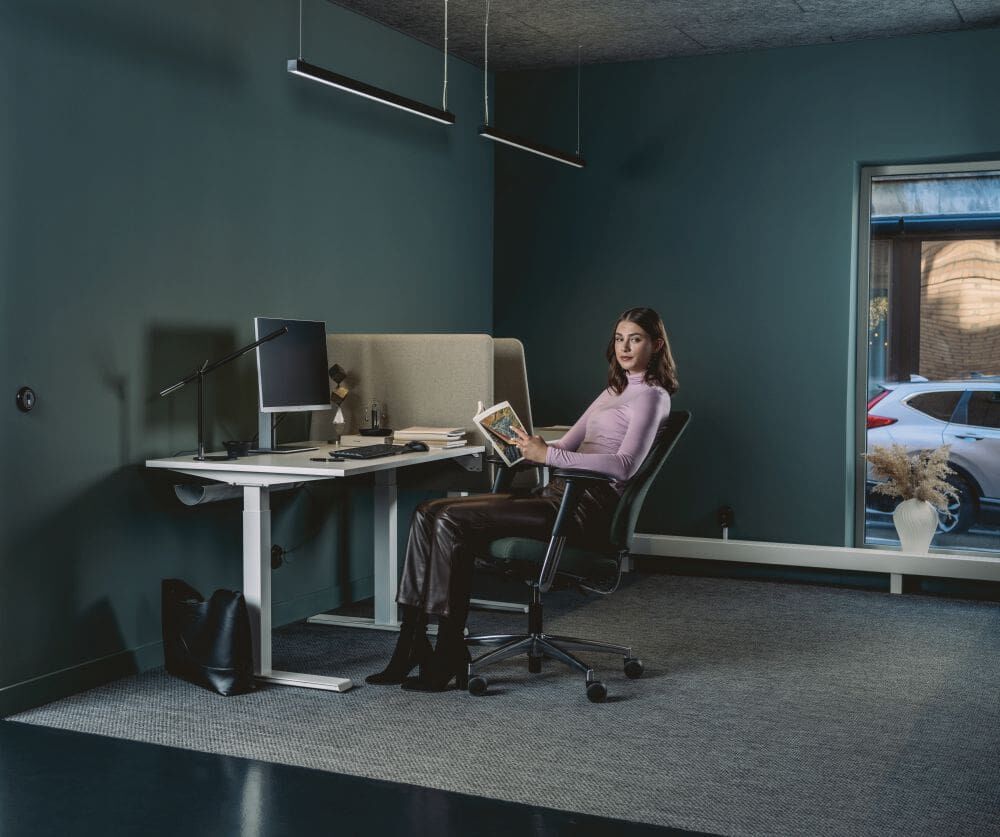 Frau sitzt auf ergonomischem Bürostuhl, hält ein Buch in modernem Büro mit stilvollem Schreibtisch und Stuhl.