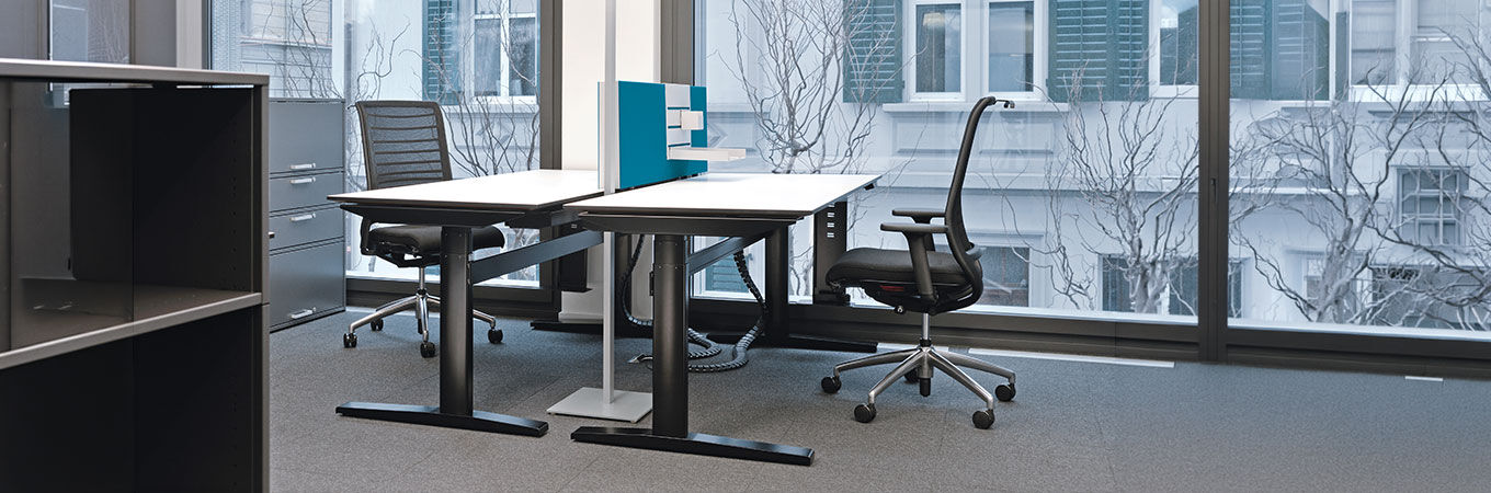Moderner Büroarbeitsplatz mit verstellbarem Schreibtisch und ergonomischen Bürostühlen vor großen Fenstern.