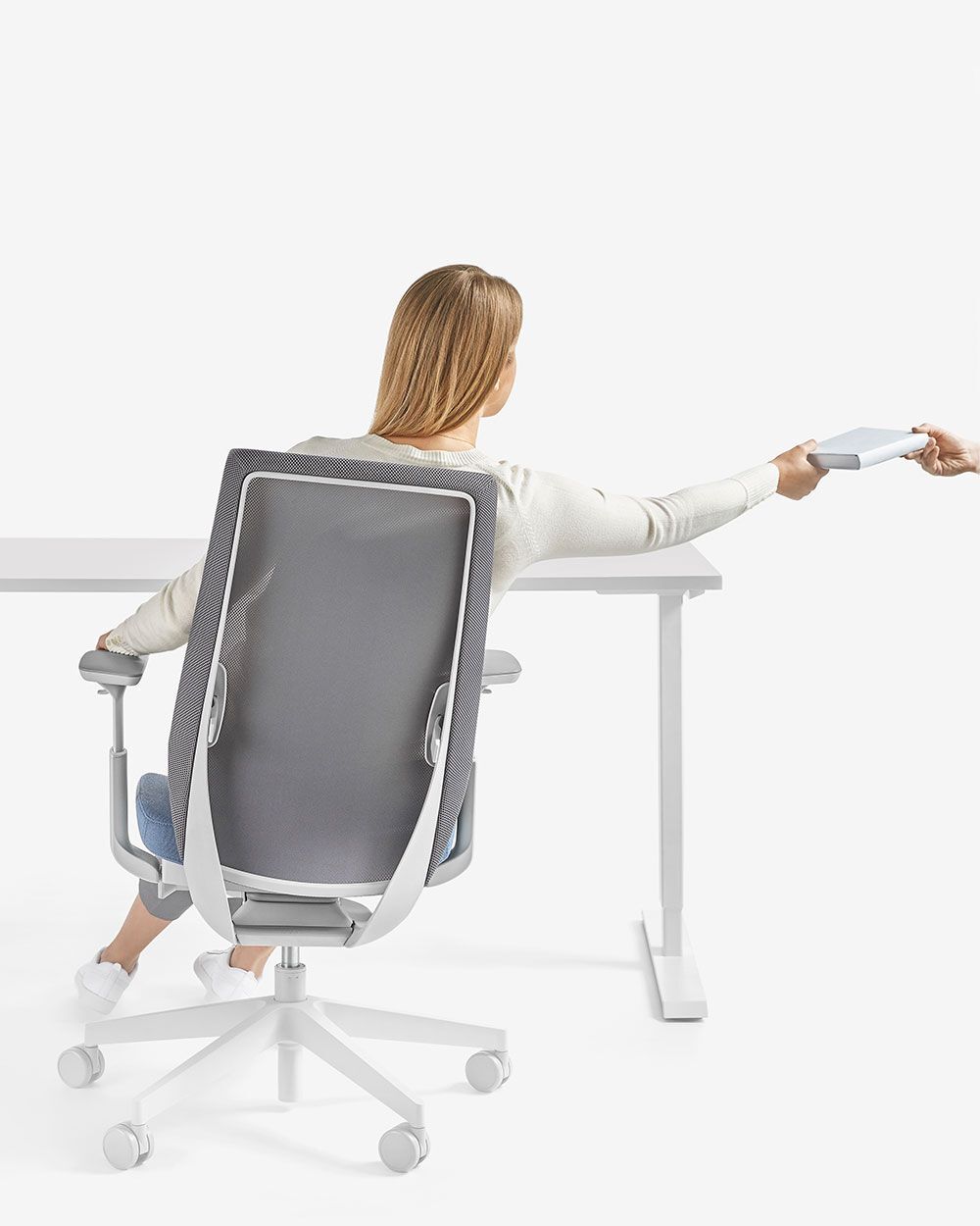 Frau sitzt auf ergonomischem Stuhl und reicht eine Decke über Schreibtisch; Betonung auf dynamischem Sitzen.
