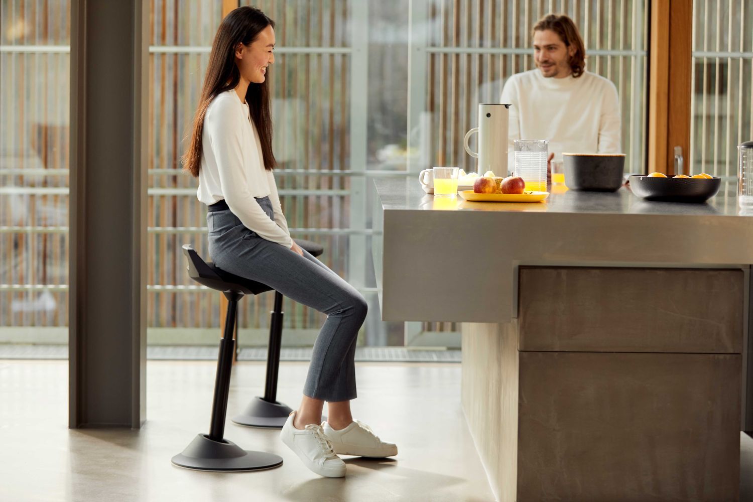 Frau sitzt auf ergonomischem Hocker an Küchentheke und spricht mit einem Mann, der steht. Moderne Inneneinrichtung.