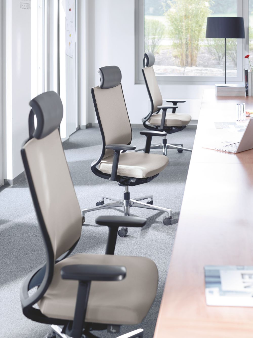 Drei ergonomische Bürostühle mit Kopfstützen in einem modernen, lichtdurchfluteten Büro neben einem Schreibtisch.