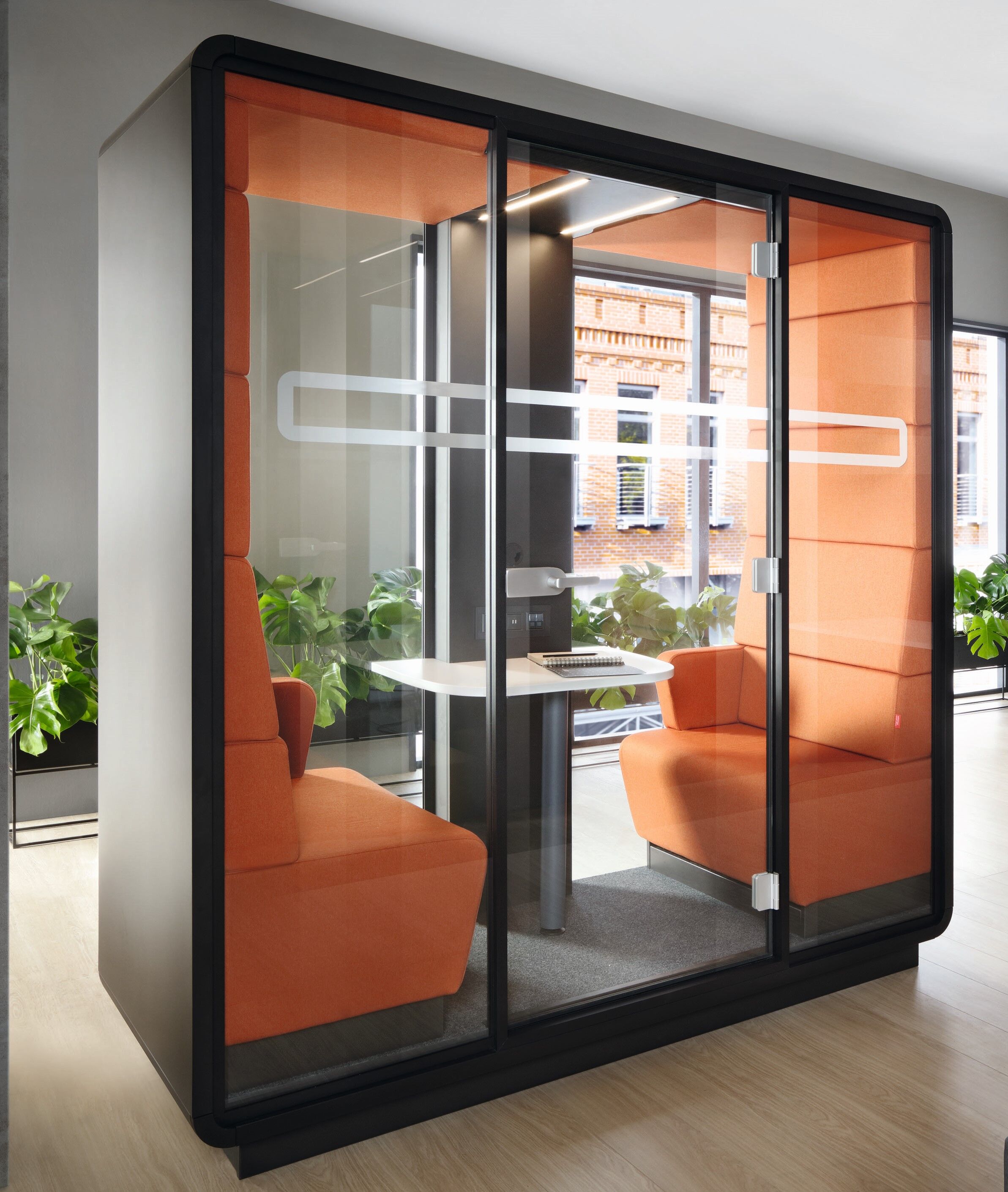 Moderne Meetingbox mit Glaswänden, zwei orangefarbenen Sitzen und Tisch, umgeben von Pflanzen, fördert konzentriertes Arbeiten.