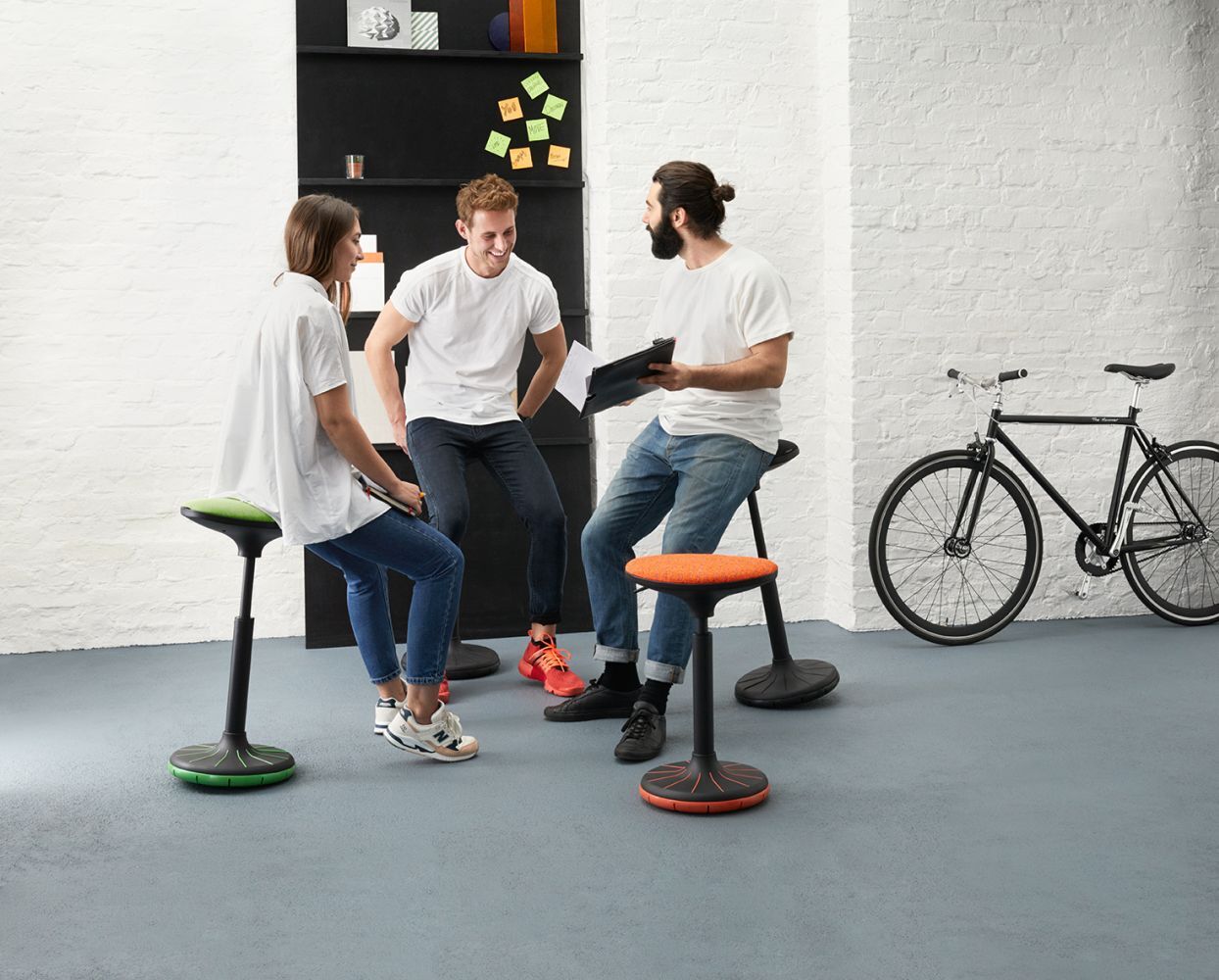Drei Personen sitzen auf ergonomischen Hockern und unterhalten sich, ein Fahrrad steht neben einer weißen Wand.