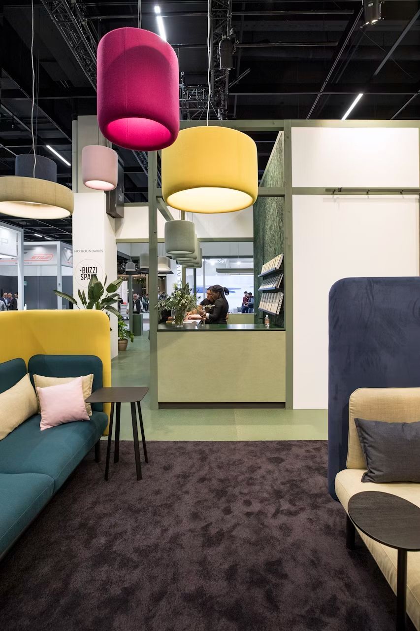 Moderne Messeausstellung mit bunten, zylindrischen Deckenleuchten und stilvollen Sitzgelegenheiten vor einer Rezeption.