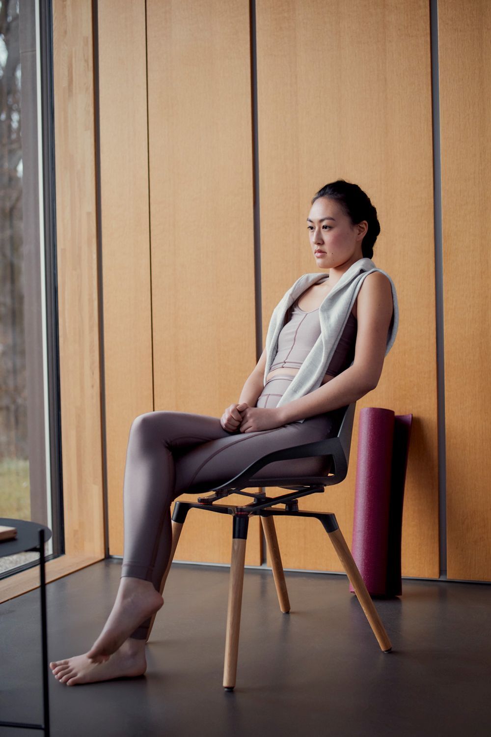 Frau sitzt entspannt auf ergonomischem Stuhl vor Holzverkleidung, mit Yoga-Matte im Hintergrund, barfuß.