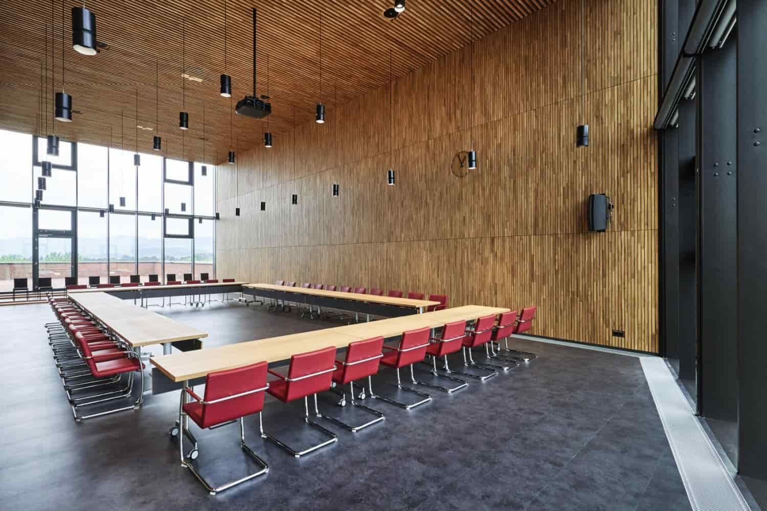 Moderner Konferenzraum mit U-förmigem Tisch und roten, ergonomischen Bürostühlen vor holzverkleideter Wand.