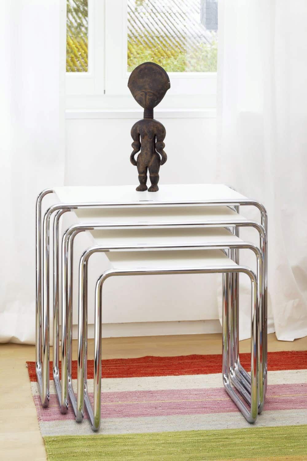 Mehrere verchromte Beistelltische, gestapelt, mit einer kleinen Skulptur darauf, vor einem Fenster.