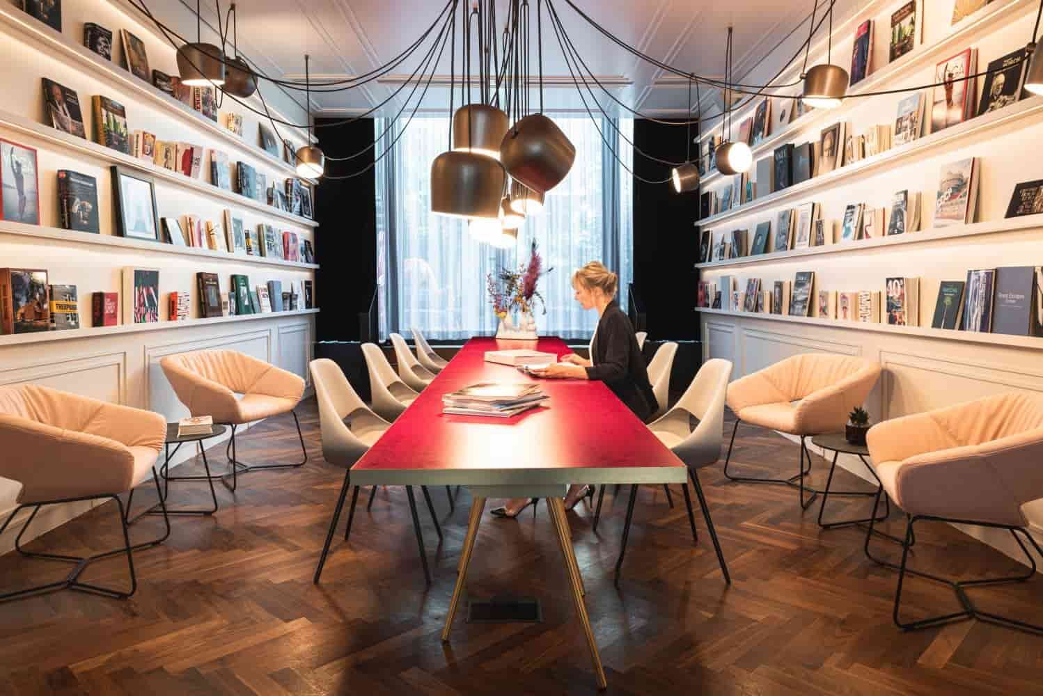 Elegantes Sitzungszimmer mit roten Tisch, modernen Stühlen und Bücherregalen, beleuchtet mit stilvollen Hängelampen.