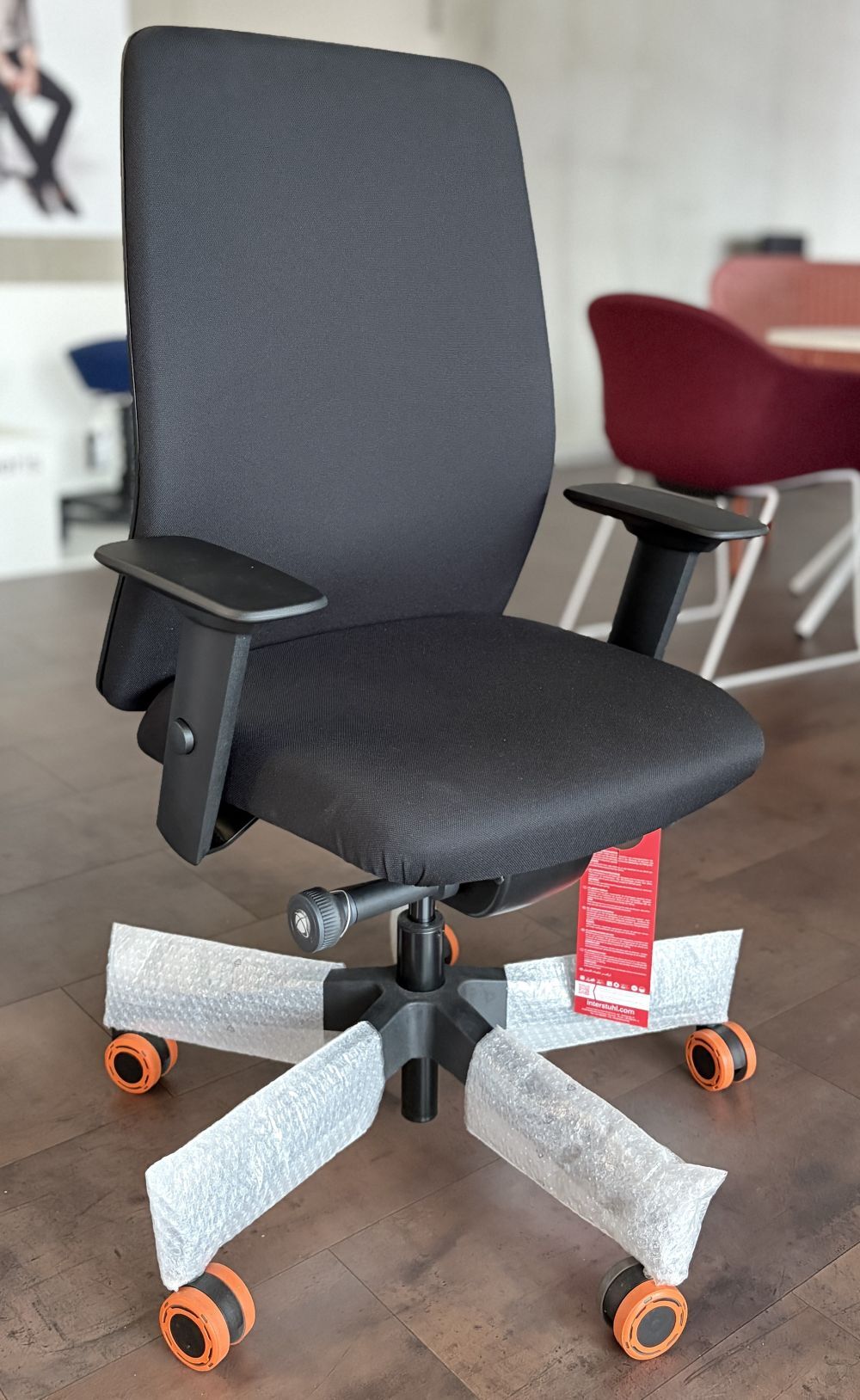 Ergonomischer Bürostuhl mit schwarzem Bezug, höhenverstellbaren Armlehnen und rollen, teils noch verpackt.