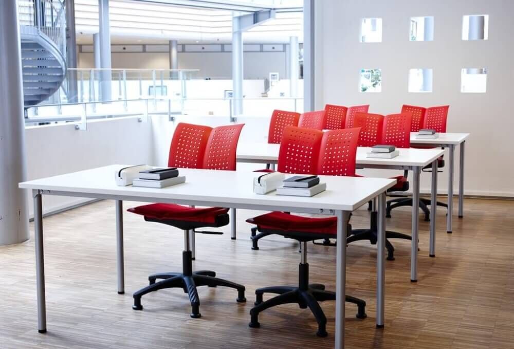 Moderne Büromöbel mit weißen Tischen und roten ergonomischen Stühlen in einem hellen Raum.