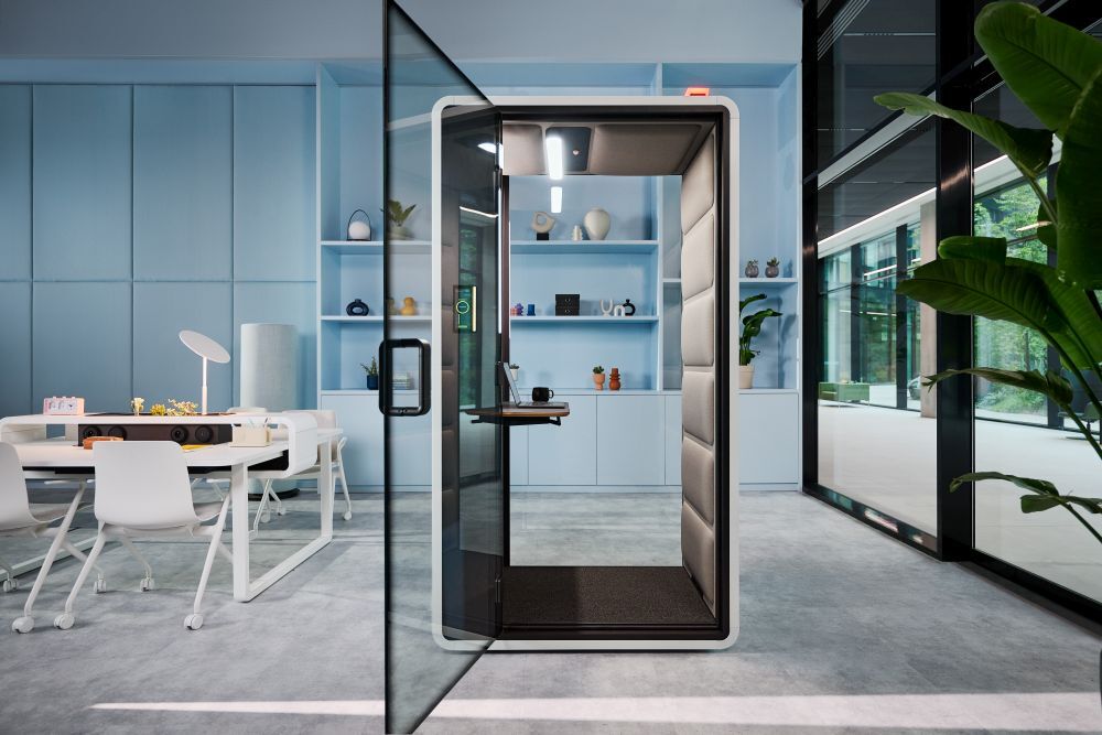 Moderne Bürokabine in offenem Raum mit Tisch, Stühlen und Pflanzen, blaues Regal im Hintergrund.