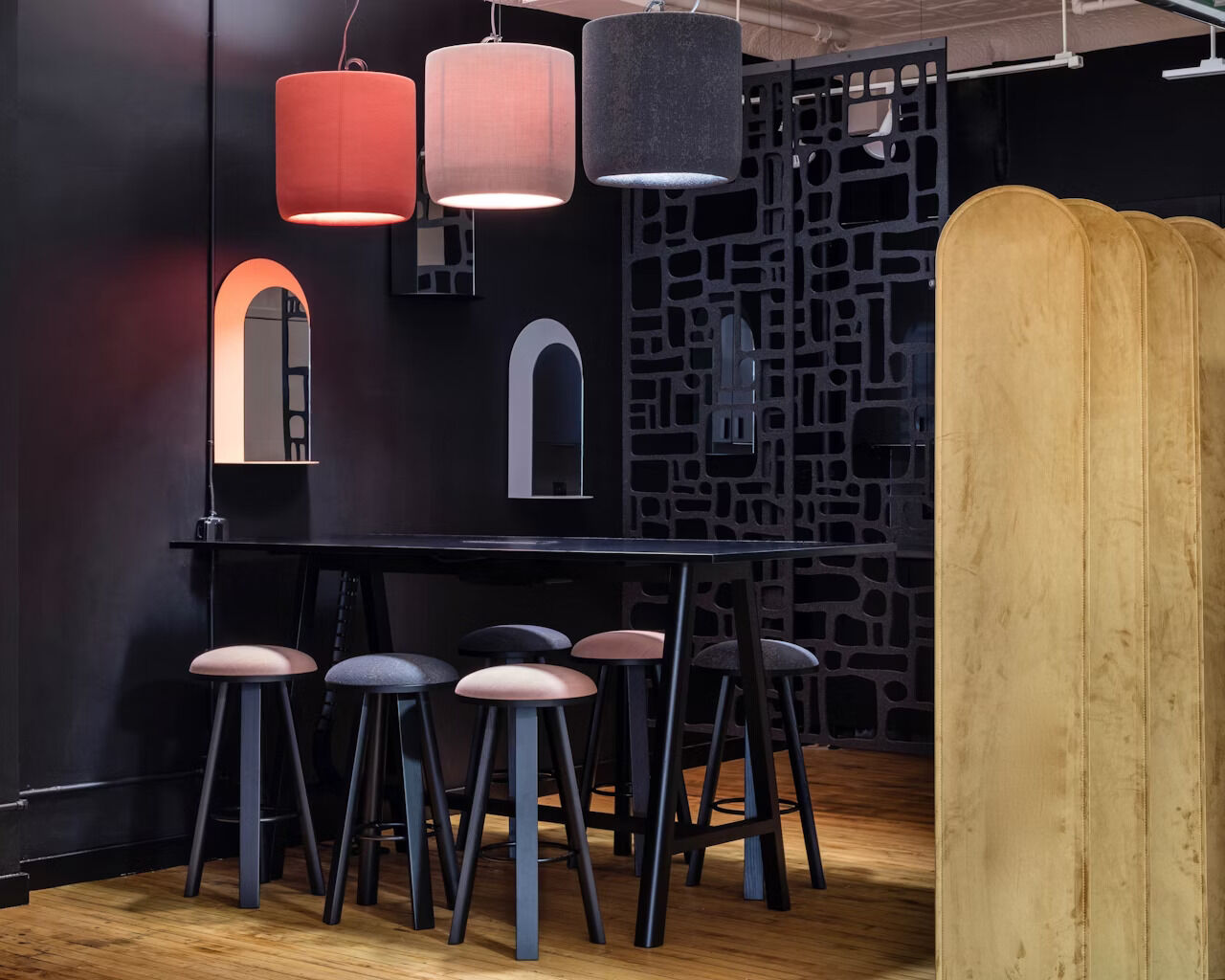 Moderner Besprechungsraum mit hohen Hockern, Tisch und dekorativen Lampen vor einer dunklen Trennwand.