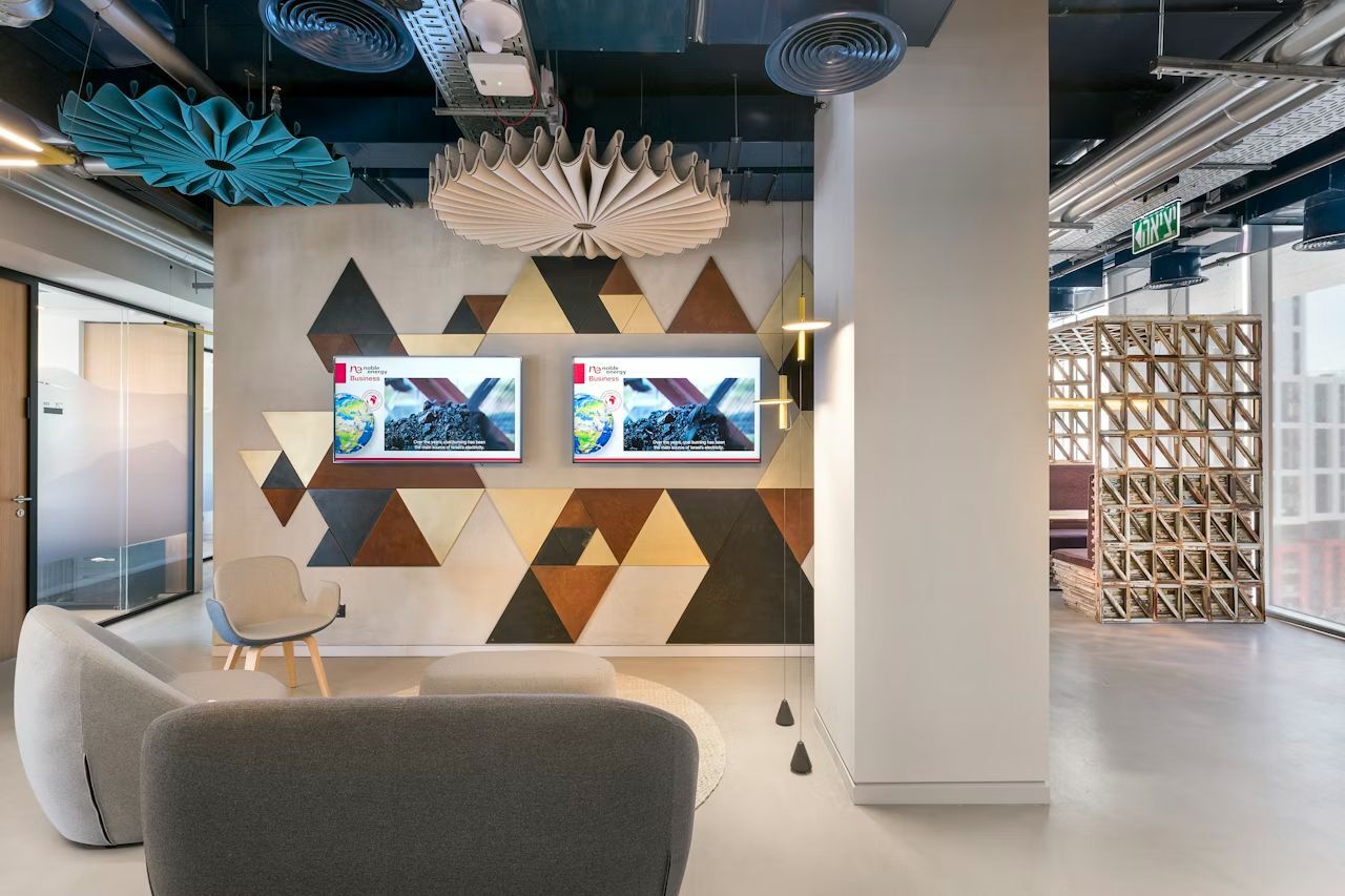 Modernes Bürointerieur mit ergonomischen Sesseln, geometrischer Wandkunst und zwei Bildschirmen für Präsentationen.