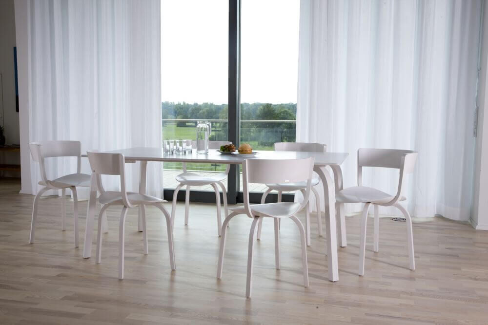 Moderner Essbereich mit sechs weißen Stühlen um einen runden Tisch vor großen Fenstern mit Landschaftsblick.