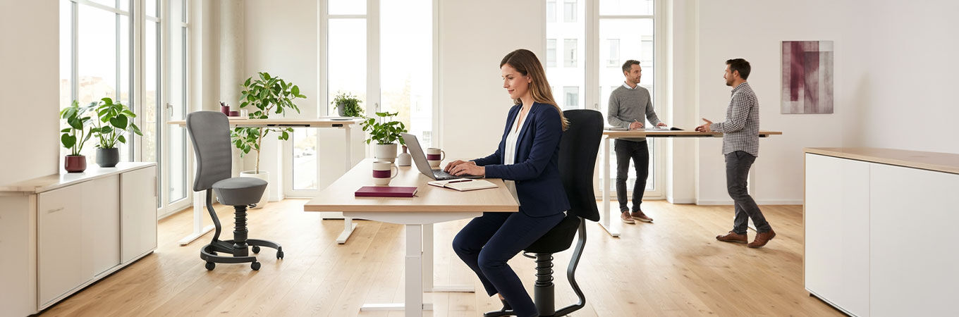 Modernes Büro mit ergonomischen Stühlen, dynamisches Sitzen, Menschen an Schreibtischen, große Fenster und Pflanzen.