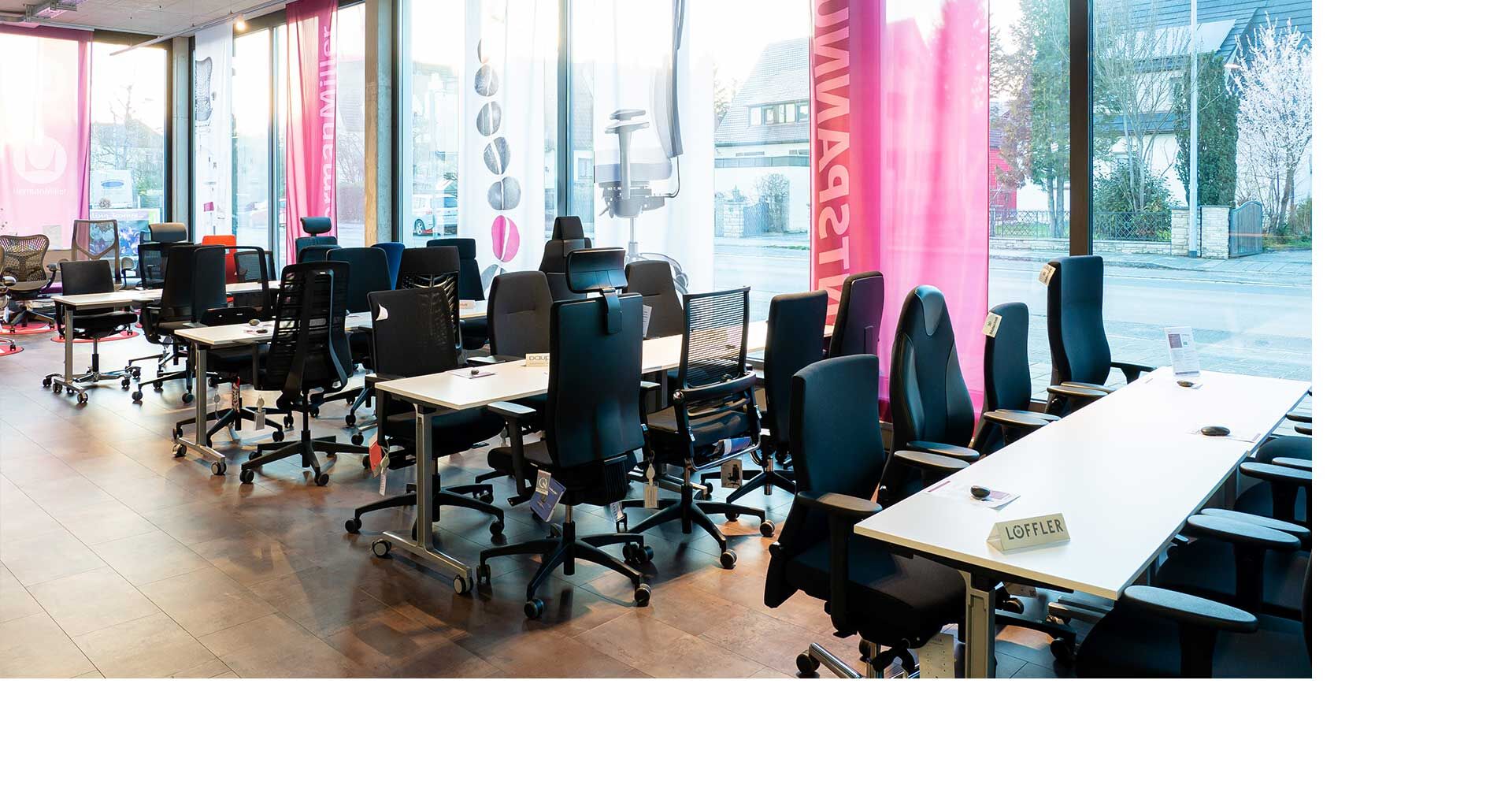 Showroom mit stilvollen ergonomischen Bürostühlen und Tischen, vor großen Fenstern mit Stadtblick.