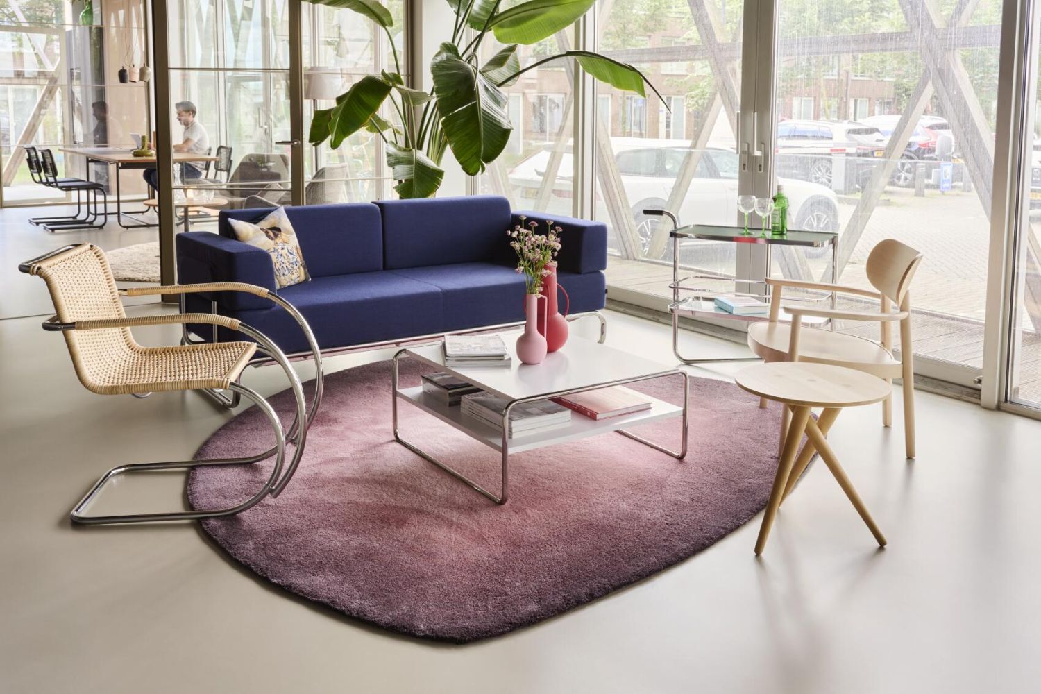 Modernes Wohnzimmer mit blauer Couch, lila Teppich, Designerstühlen und großen Zimmerpflanzen vor einer Glasfront.