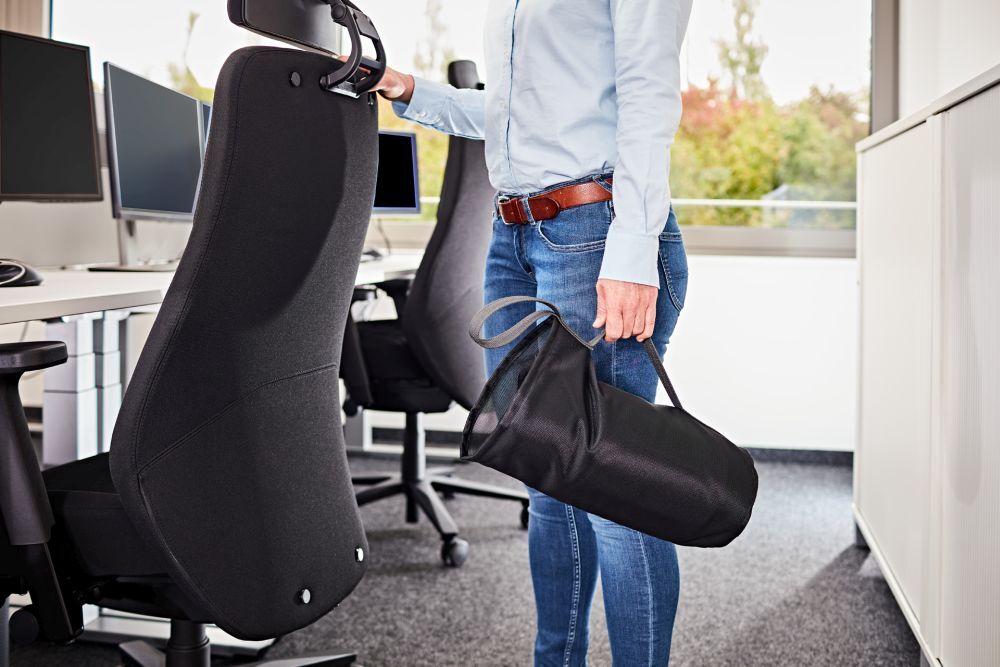 Person im Büro mit ergonomischem Stuhl, hält schwarze Tragetasche, zeigt dynamisches Sitzen.