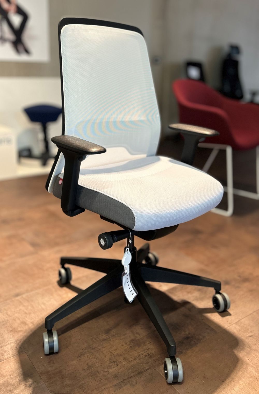 Ergonomischer Bürostuhl mit Netzrückenlehne und Rollen, auf einem Holzboden in einem Showroom ausgestellt.