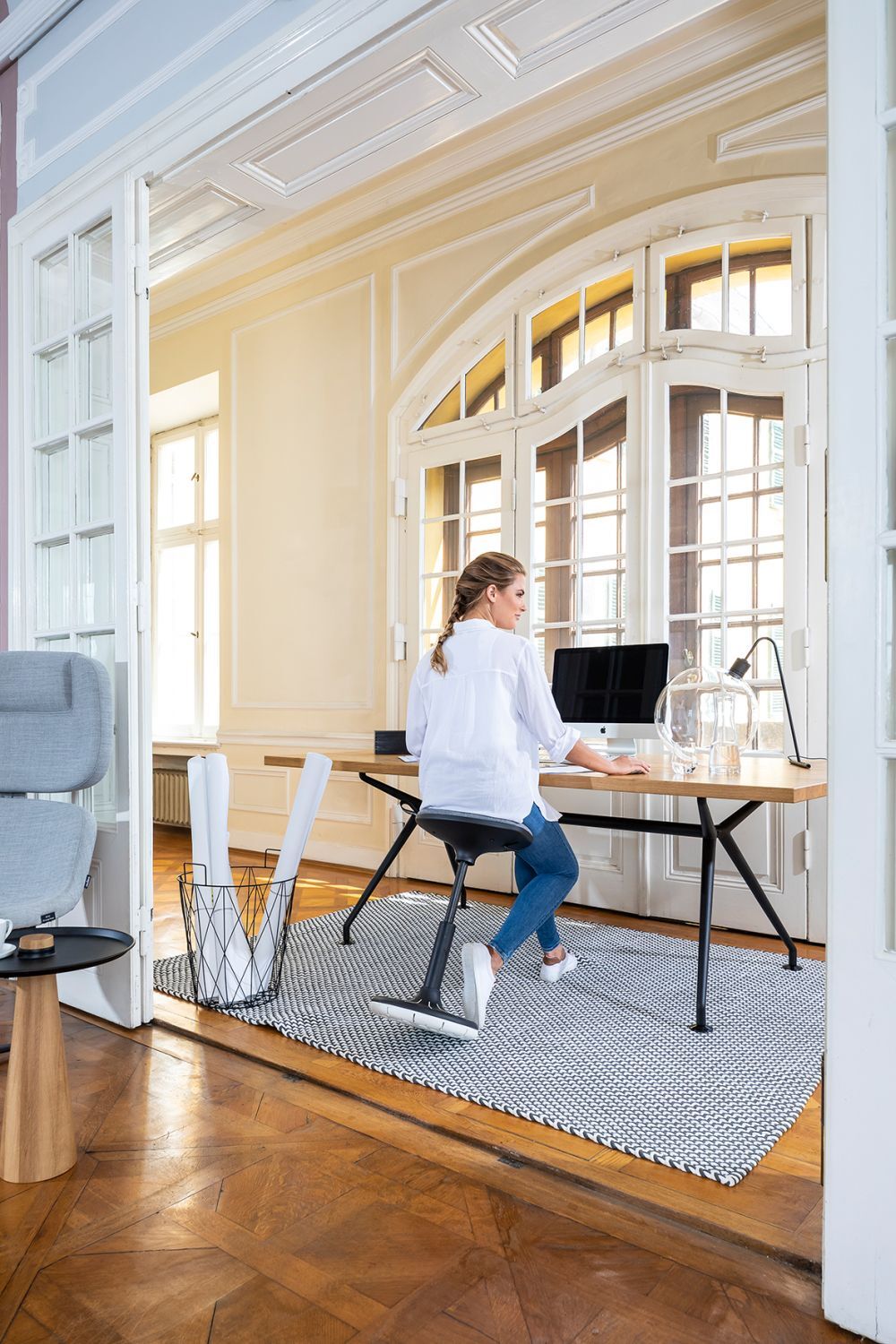 Frau sitzt auf ergonomischem Stuhl am Schreibtisch in hellem, eleganten Arbeitszimmer mit Holzboden.