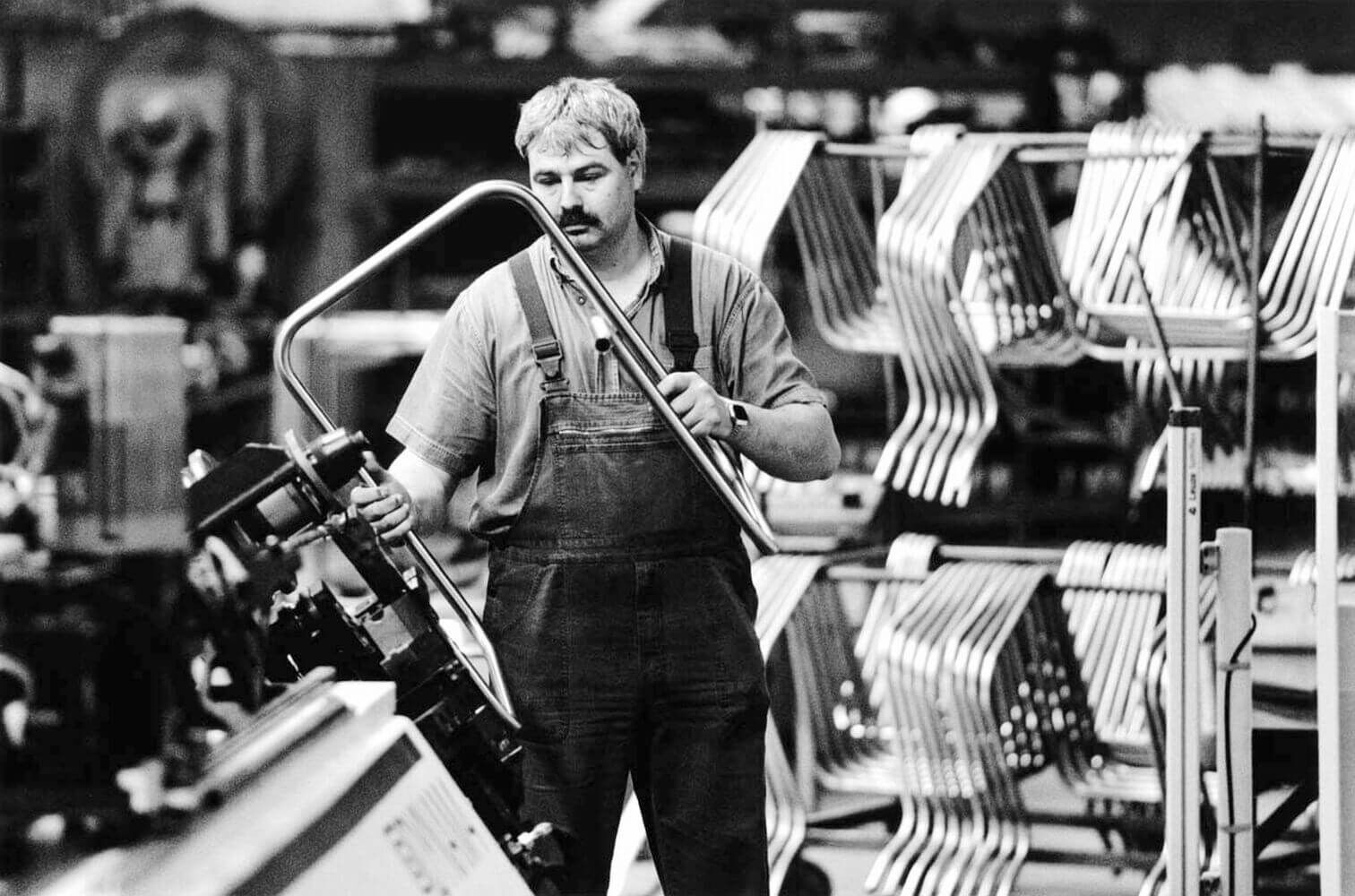 Arbeiter in einer Werkstatt, der ein Metallrohr formt; im Hintergrund gestapelte Metallrahmen.