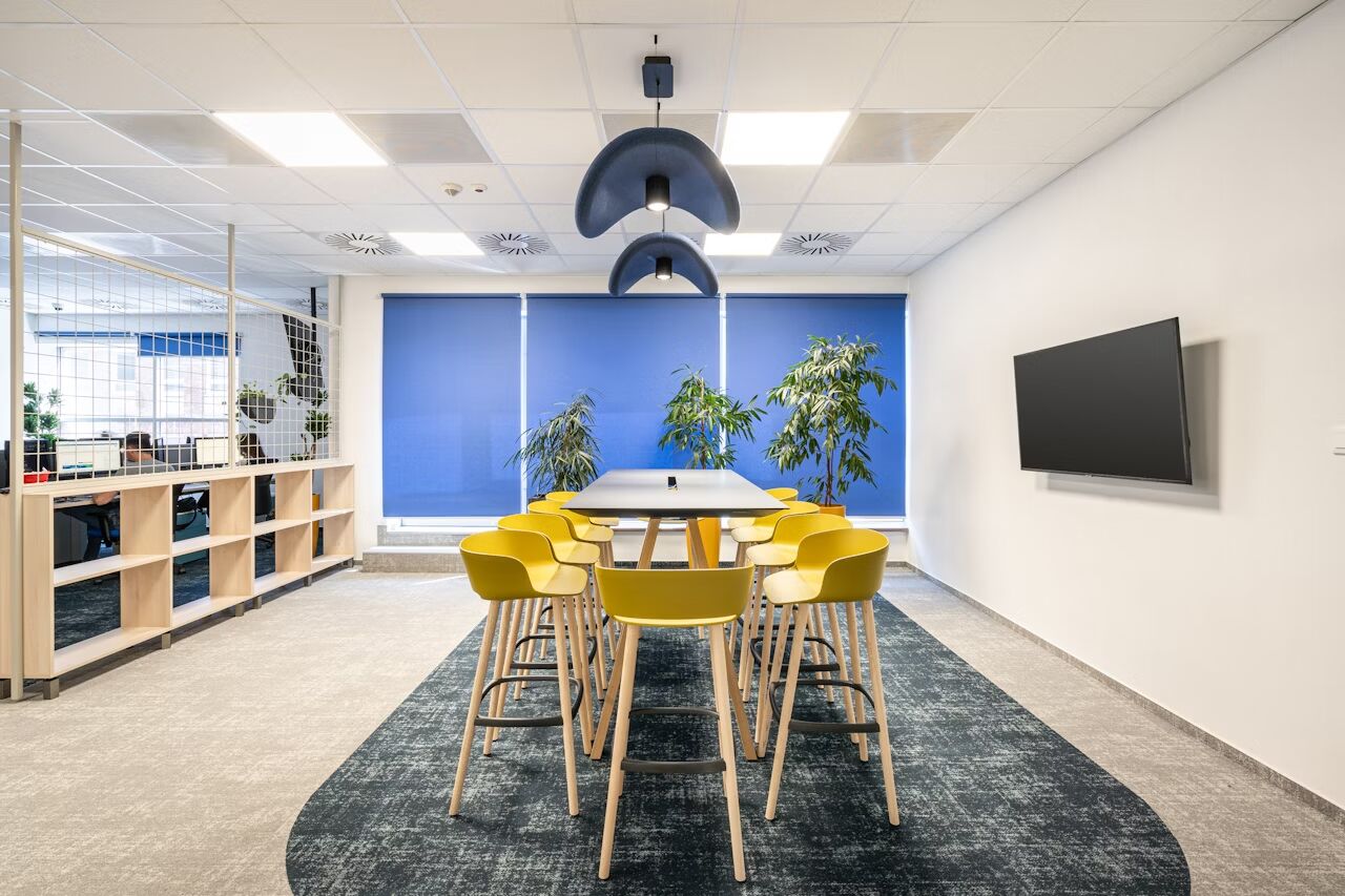 Modernes Besprechungszimmer mit gelben Stühlen, großem Tisch, Hängelampen, Pflanzen, Fernseher und blauen Jalousien.