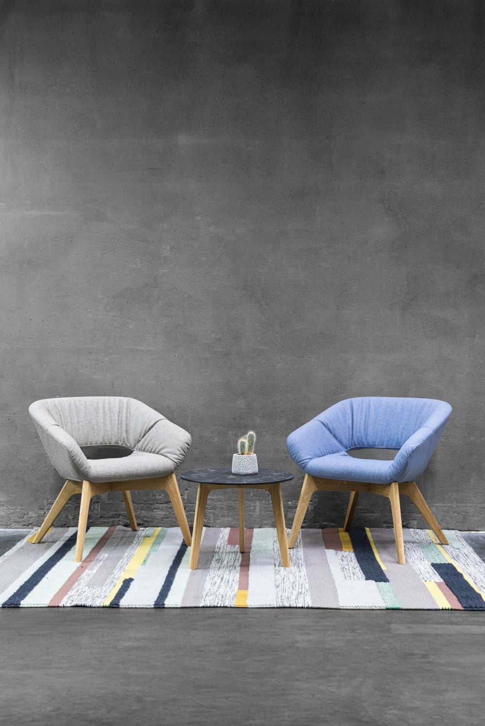 Zwei Stühle, grau und blau, mit Tisch und Kaktus auf gestreiftem Teppich vor grauer Wand.