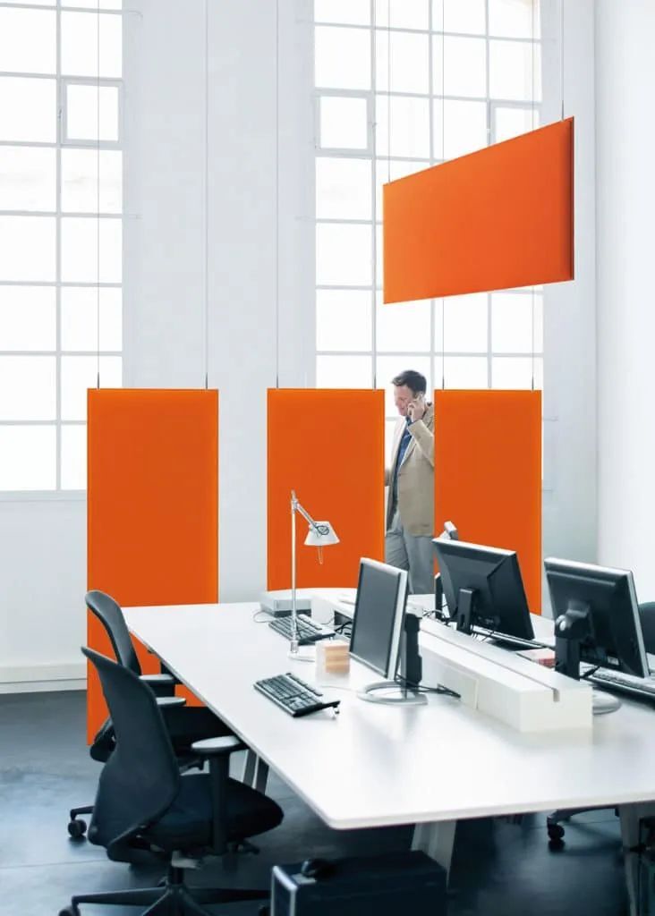 Modernes Büro mit weißen Schreibtischen, ergonomischen Stühlen und orangefarbenen Akustikpaneelen. Person telefoniert.