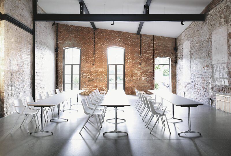 Heller Konferenzraum mit langen Tischen, weißen Stühlen und Ziegelwänden, ideal für Meetings und Zusammenarbeit.