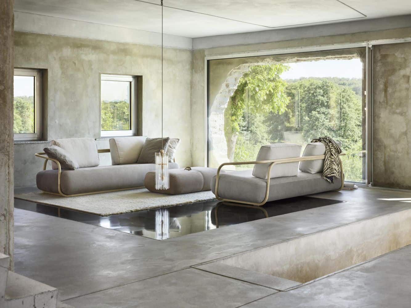 Modernes Wohnzimmer mit grauem Sofa, großen Fenstern und Blick auf einen grünen Wald im Hintergrund.
